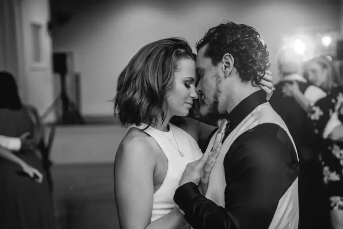 A couple shares an intimate moment while slow dancing at an indoor event, surrounded by other people in the background.