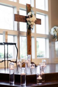 20180415_SorayaA_W-0404-600×900-1 A wooden cross decorated with flowers stands on a platform, surrounded by tall glass vases with floating candles. Large windows provide natural light in the background.