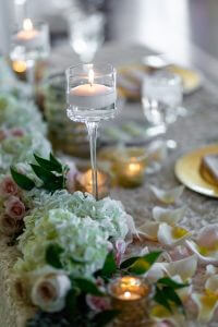 20180415_SorayaA_W-0422-600×900-1 A close-up of an elegant table setting with a lit candle in a glass holder, white flowers, rose petals, and glassware on a white tablecloth.