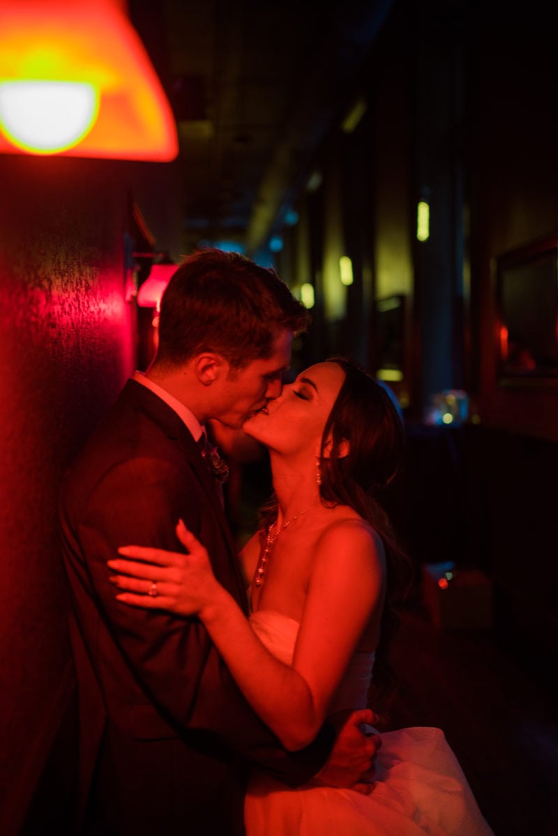 A bride and groom share a passionate kiss in a dimly lit hallway with red and yellow lighting.