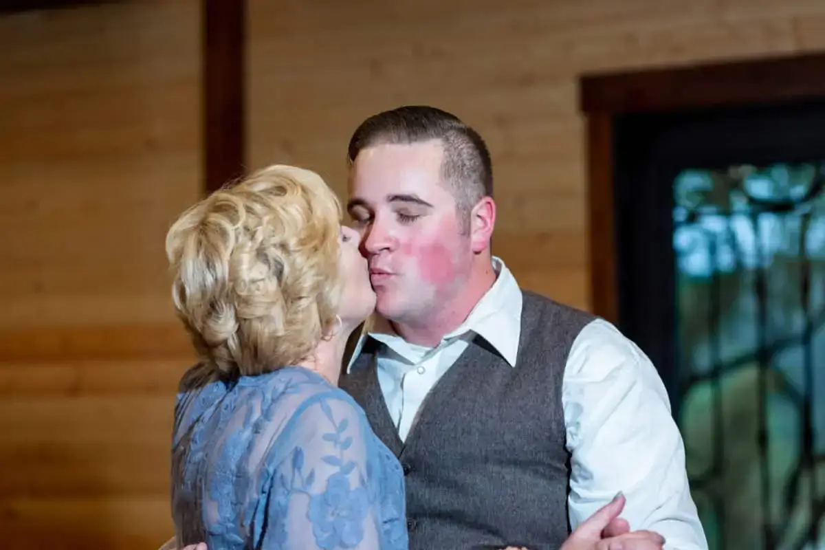 A woman in a blue lace dress and a man in a gray vest and white shirt share a kiss indoors against a wooden wall background.