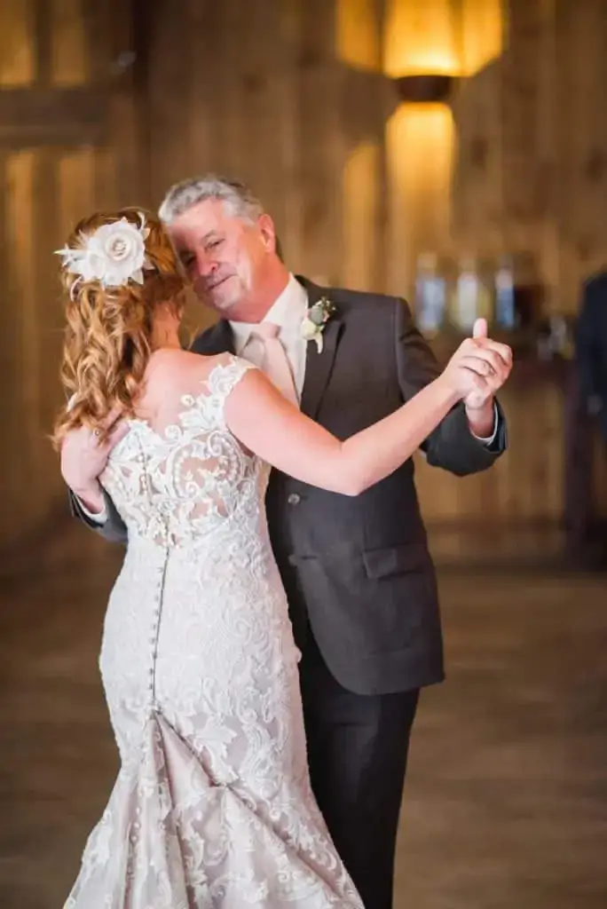 A bride in a lace wedding dress dances with an older man in a suit in a warmly lit, wooden interior.