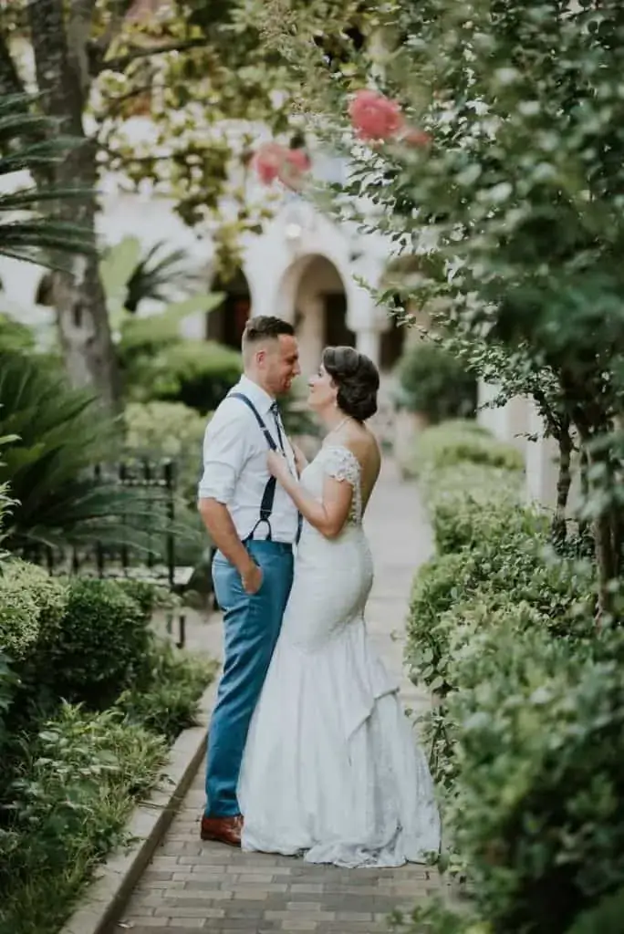A bride and groom stand on a garden pathway facing each other, surrounded by greenery and soft outdoor lighting.