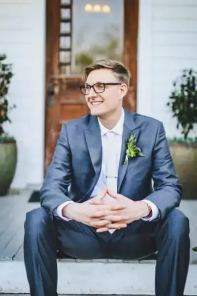 A man in a blue suit with a boutonniere sits on steps in front of a wooden door, smiling and looking to the side.