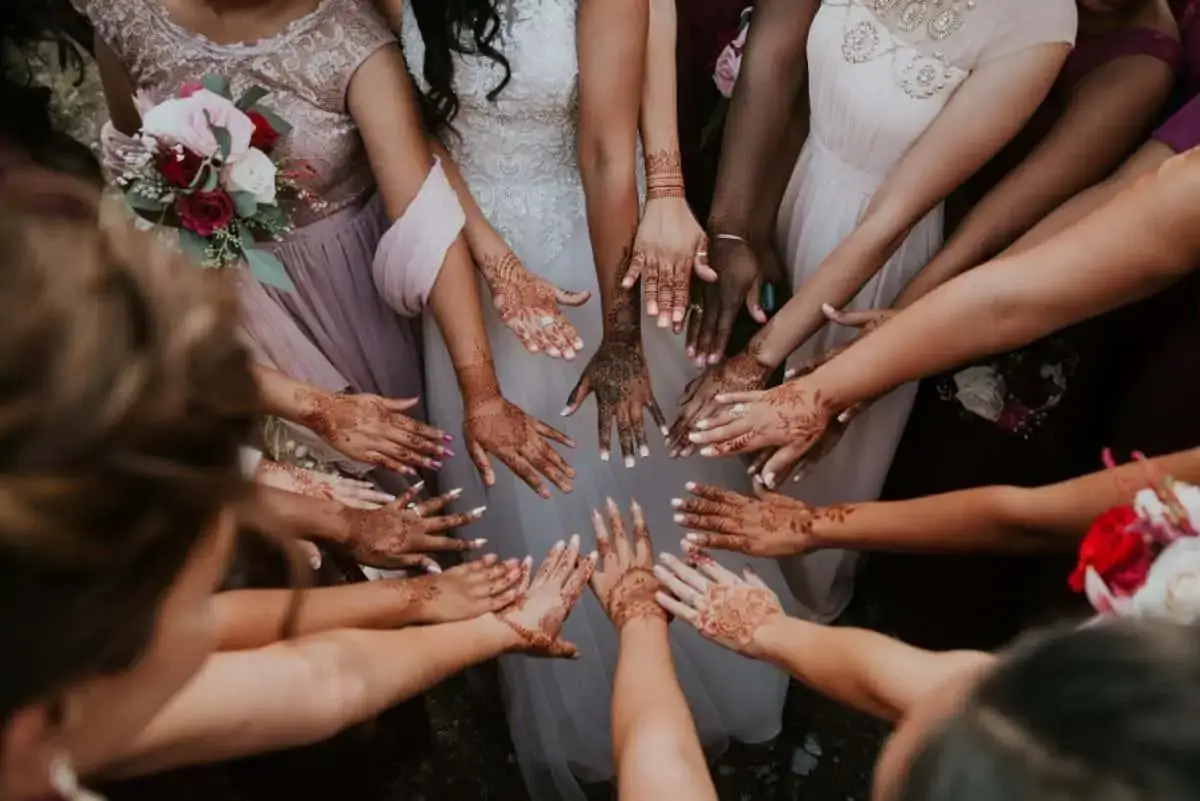 A group of women in dresses show their hands decorated with henna designs, arranged in a circle around a bride in a white gown.