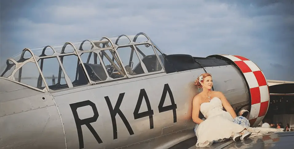 A woman in a white wedding dress sits on the wing of a silver aircraft with the markings "RK44" and a red-and-white checked tail.