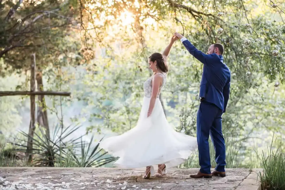 A bride in a white dress twirls while holding hands with a groom in a blue suit on a stone path surrounded by greenery at sunset.