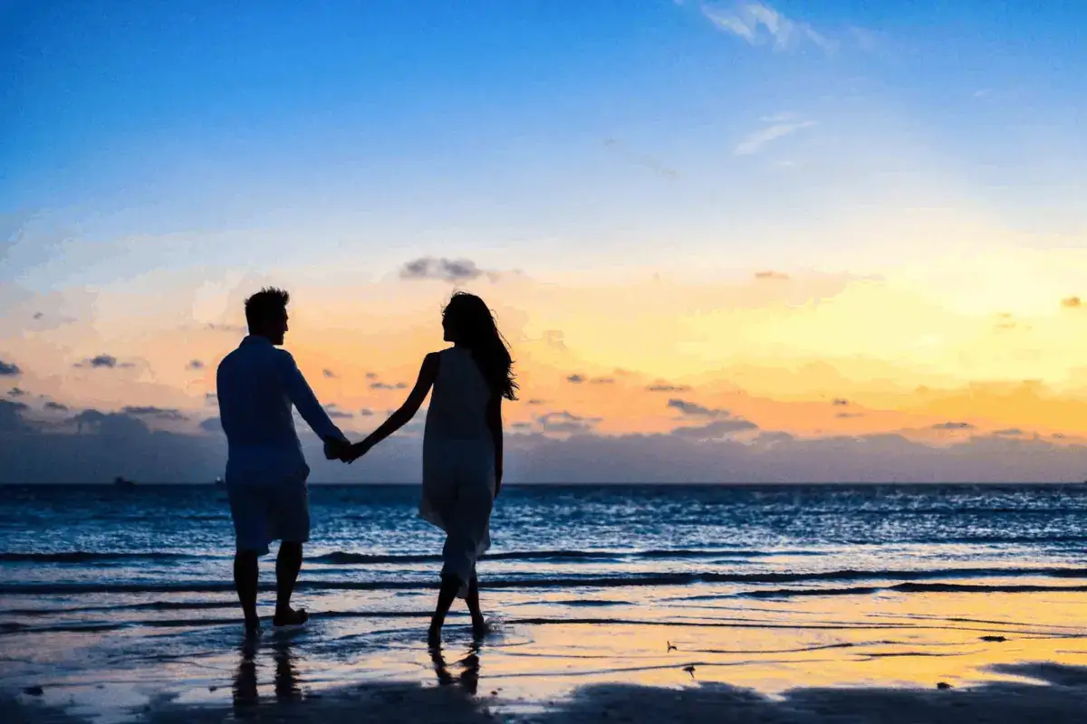 A couple holds hands while walking along the shoreline at sunset, with the sky displaying shades of blue, orange, and yellow.