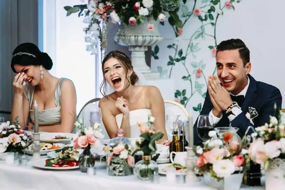 Three people in formal attire sit at a decorated table, laughing and smiling during what appears to be a wedding celebration.