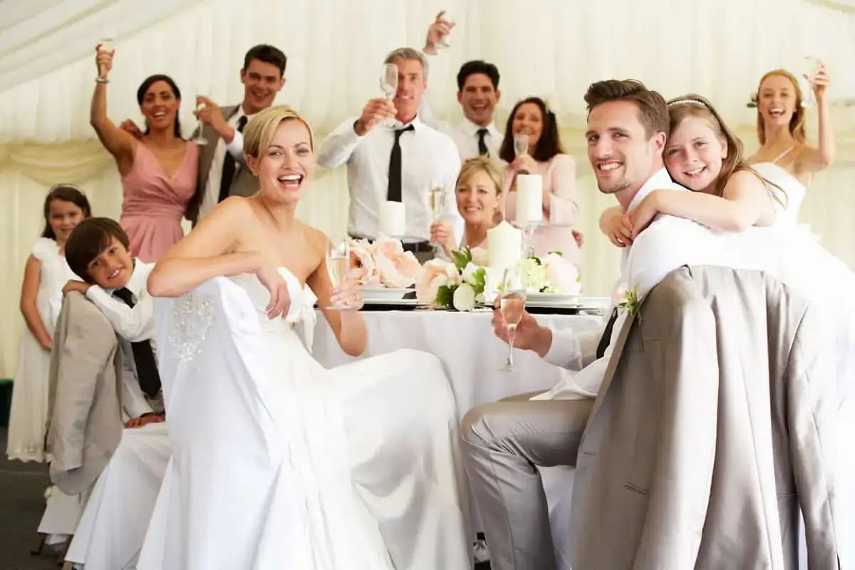 A group of people dressed formally pose and smile for a photo at a wedding reception, raising glasses in a celebratory toast around a decorated table.