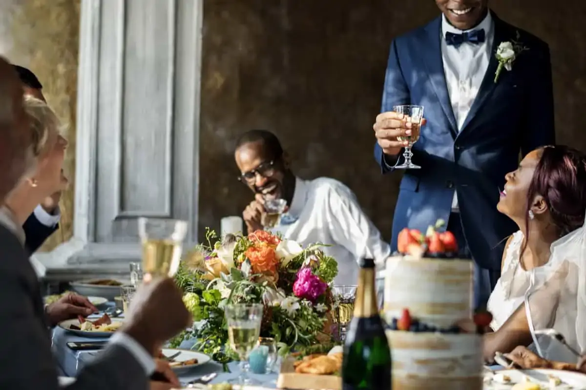 Five people sit around a decorated table with flowers and a cake, smiling and holding glasses, as one person in a suit stands and gives a toast.