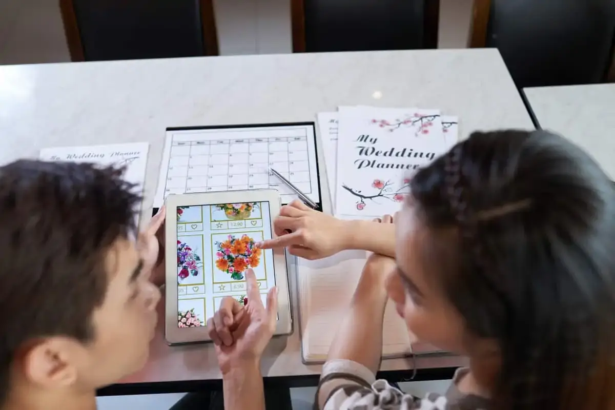 Two people look at a tablet displaying floral arrangements, with wedding planning documents and a calendar on the table.