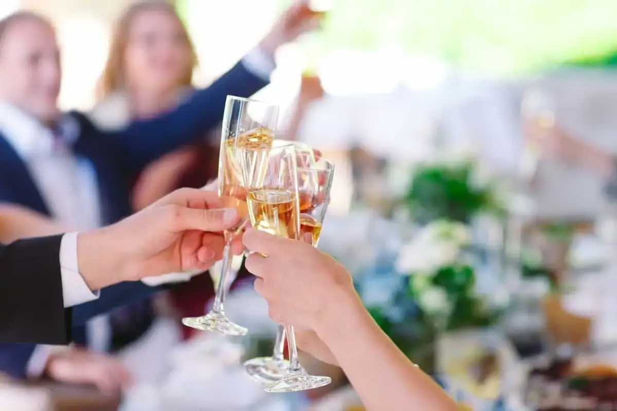 Several people hold and clink champagne glasses in a toast at a gathering, with a decorated table and blurred figures in the background.