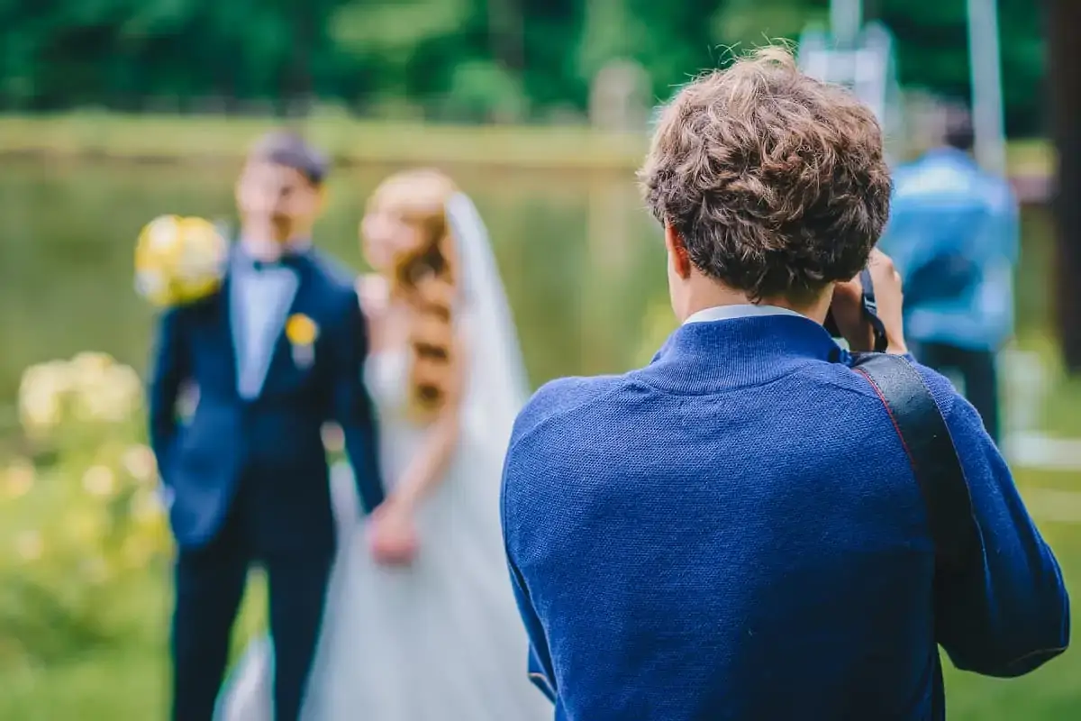 A photographer takes a picture of a bride and groom standing together outdoors near a body of water.