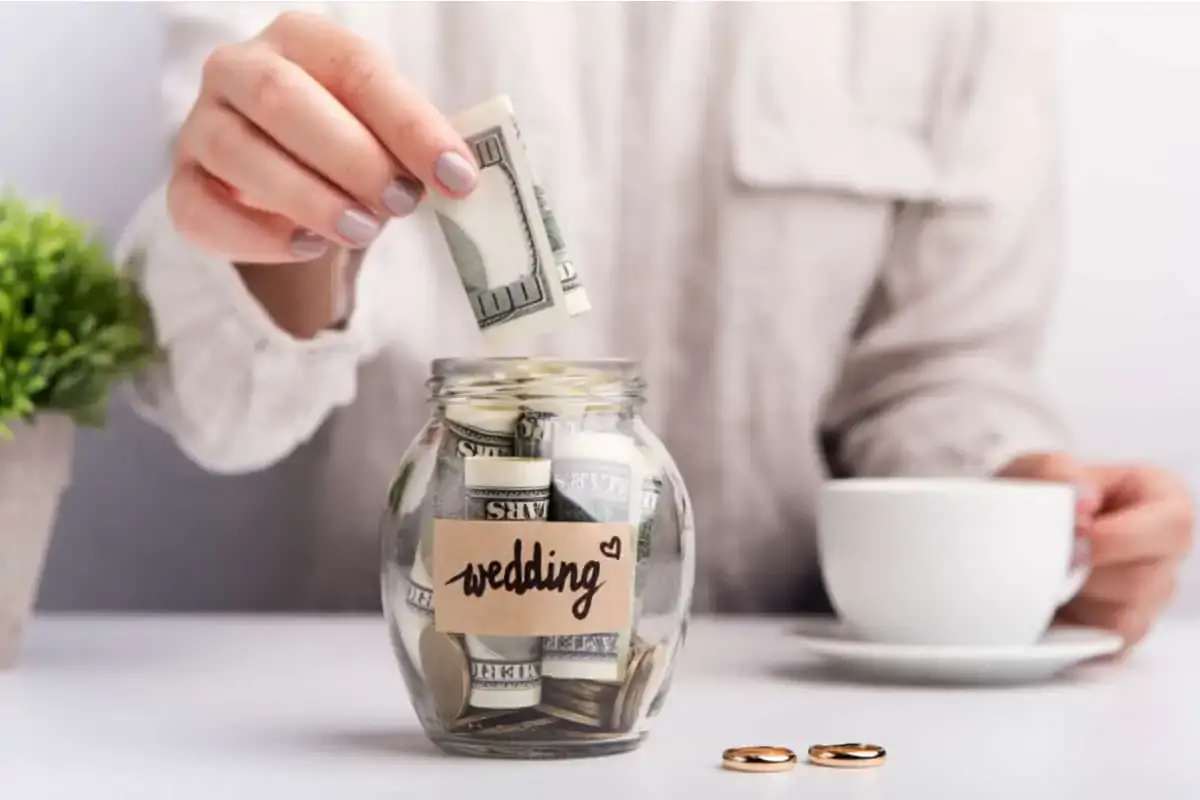 A person places a folded dollar bill into a glass jar labeled "wedding," with more cash inside; two rings and a cup are on the table.