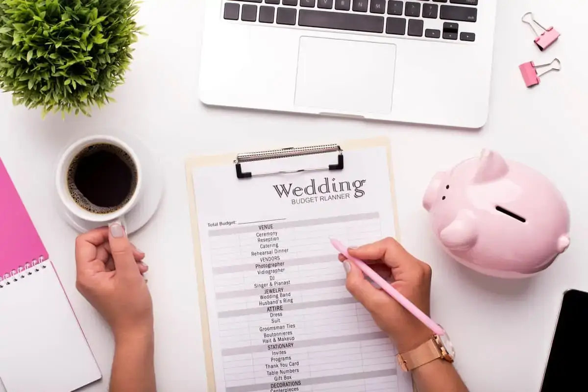 Person holding a pink pen fills out a wedding budget planner on a clipboard, with a coffee cup, laptop, plant, and pink piggy bank on the desk.