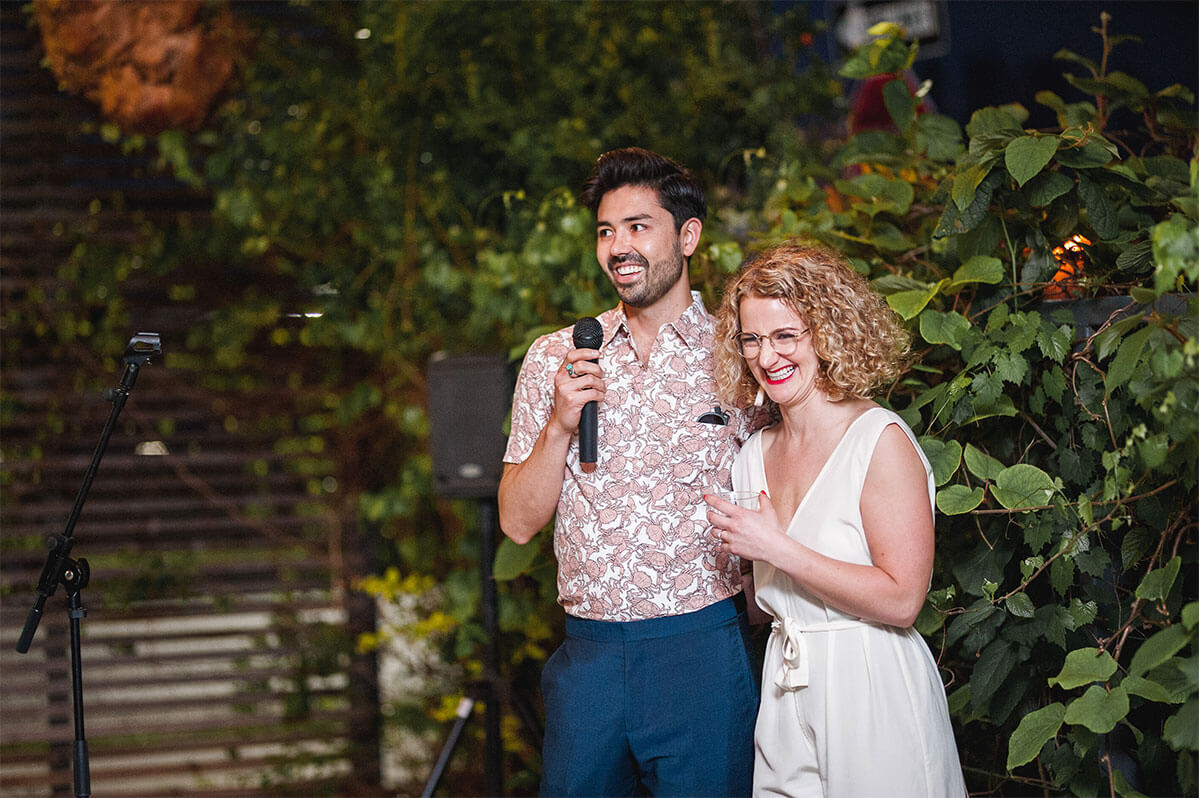 A man holding a microphone stands next to a smiling woman in a white outfit, both facing forward, with greenery in the background.