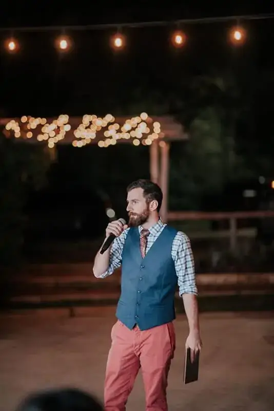 A man in a blue vest, plaid shirt, and red pants holds a microphone and a tablet, speaking outdoors at night with string lights—perfect for anyone seeking a DJ near Austin TX for wedding celebrations.