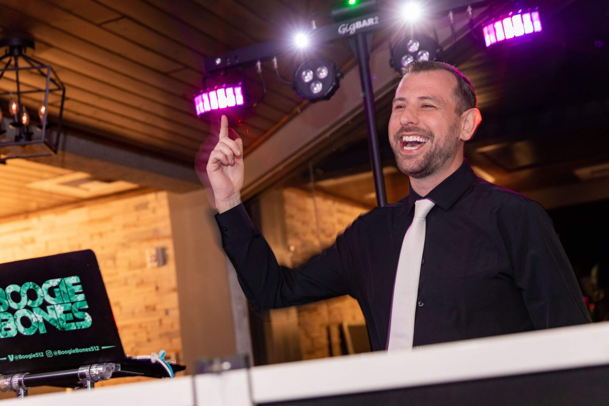 A DJ in a black shirt and white tie stands behind a booth, smiling and raising one finger amid colorful lights—a perfect example of the fun atmosphere you can expect when considering the price of a wedding DJ.