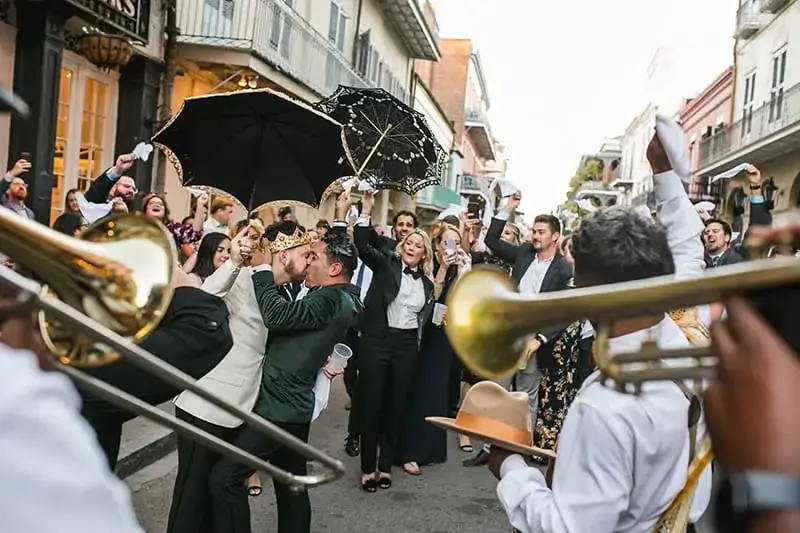 A wedding parade proceeds down a city street, featuring musicians with brass instruments, people holding umbrellas, and a joyful crowd—an ideal scene for wedding videographers in New Orleans to capture unforgettable moments.