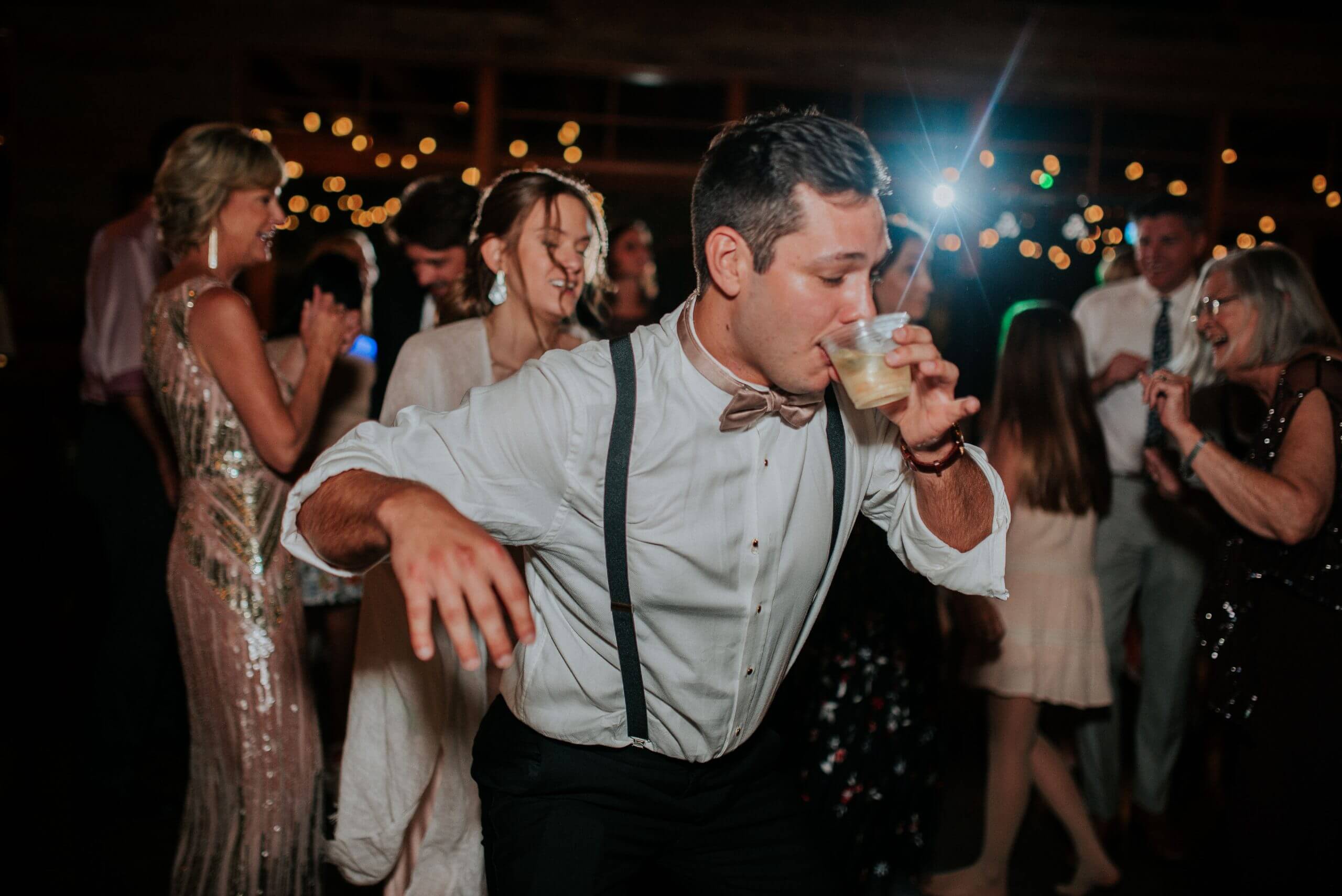 A man in formal attire drinks from a plastic cup on a crowded dance floor at a party, while a professional DJ energizes the crowd and other people dance and mingle around him.