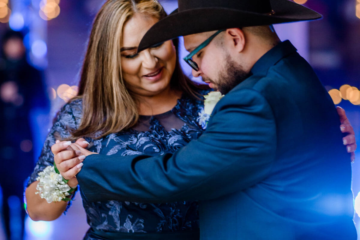 A woman in a blue dress and a man in a dark suit and cowboy hat dance together, holding hands, beneath glowing wedding uplighting, with string lights blurred in the background.