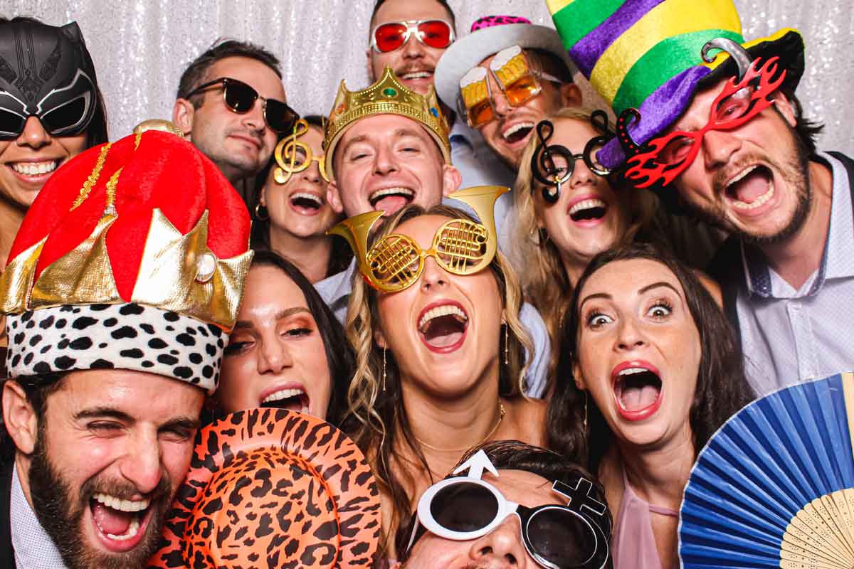 A group of people pose closely together, wearing colorful, oversized hats, novelty glasses, and holding props, smiling and laughing against a glittery backdrop—showing why knowing when to reserve photo booth fun is key for your event!.