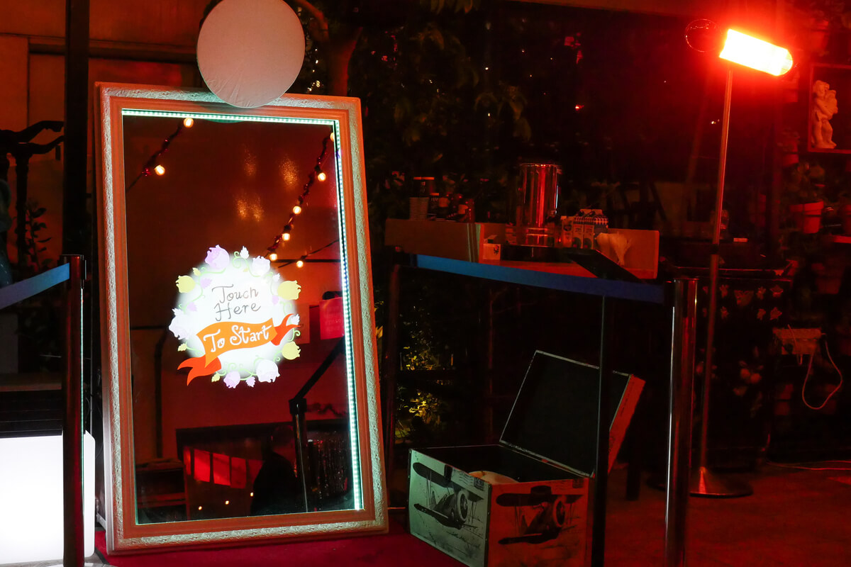 A large interactive mirror photo booth with a display reading "Touch Here to Start" is set up at an indoor event, surrounded by string lights and various equipment.