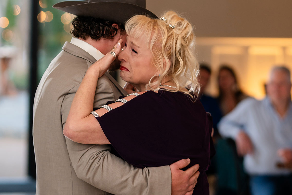 A woman wipes tears while hugging a man in a suit and cowboy hat at an indoor event, perhaps sharing an emotional moment inspired by unique mother-son dance songs, with other people blurred in the background.