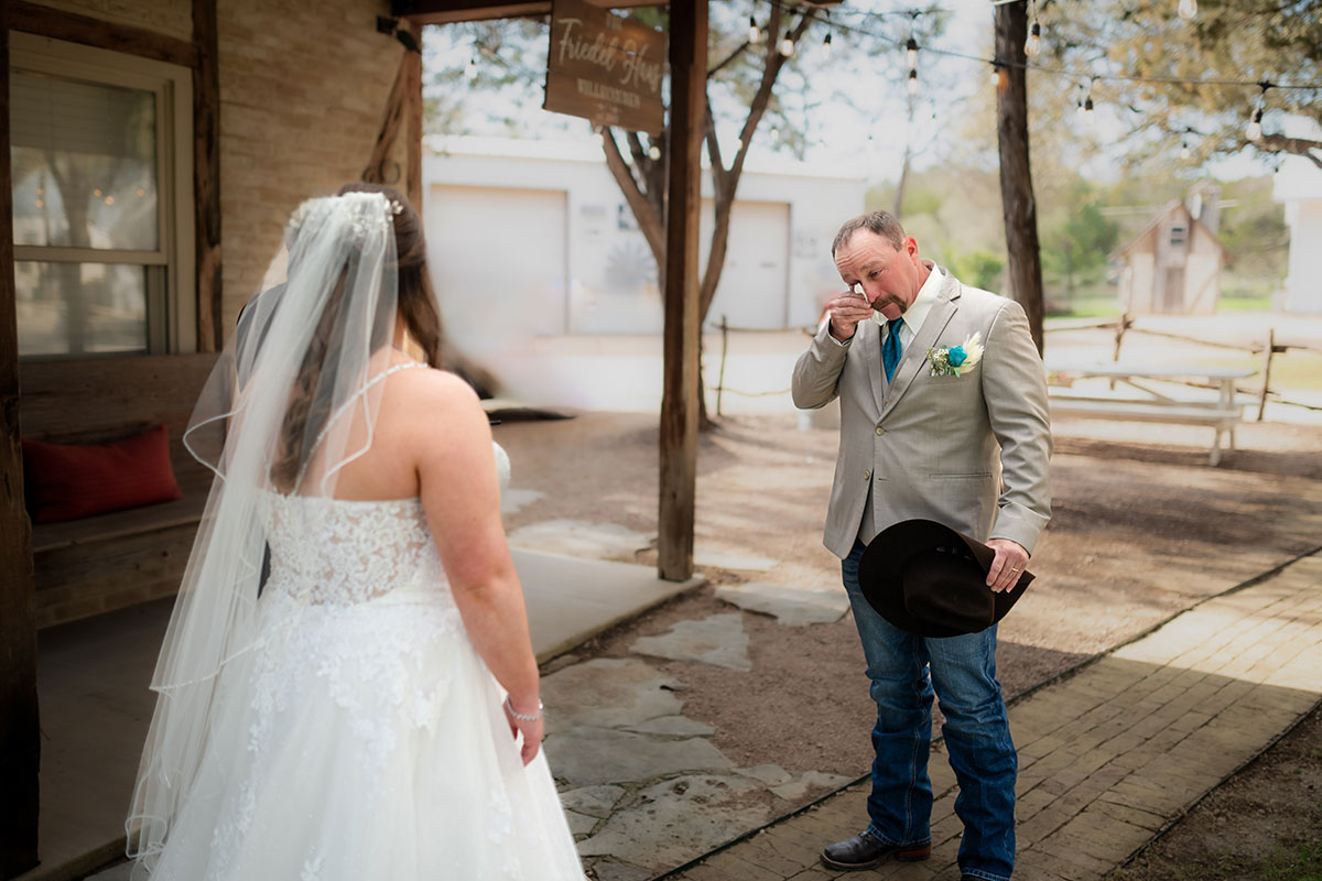 A bride in a white dress stands before a man in a beige jacket and jeans, wiping his eyes and holding a black hat, outside a rustic building—capturing the emotion found in heartfelt songs about daughters.