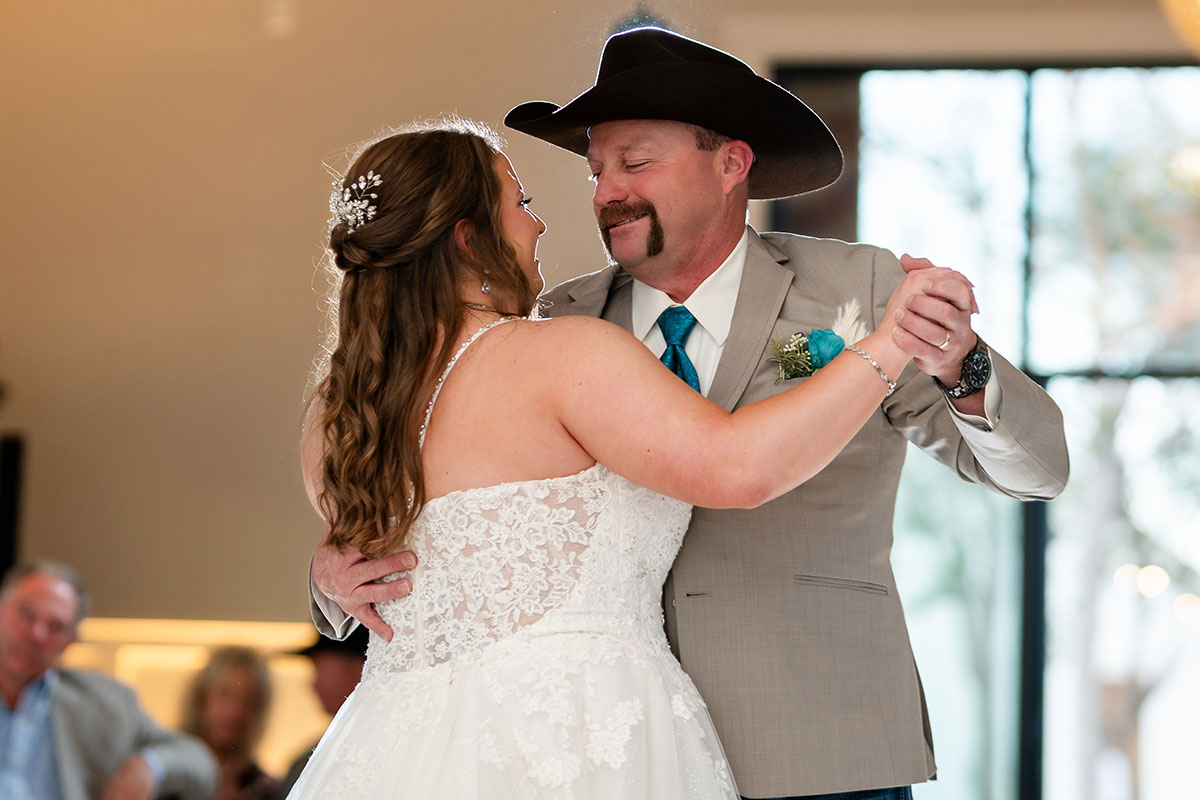 A bride in a white lace dress and a man in a tan suit with a cowboy hat share a joyful moment indoors, smiling as they dance together to one of their unique father-daughter dance songs.