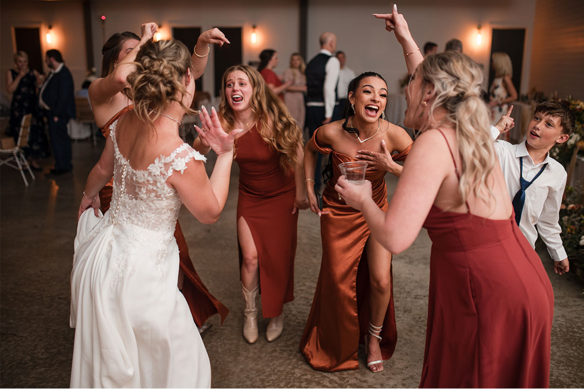 A group of people in formal attire, including a bride in a white dress, are laughing and dancing excitedly at an indoor event to the best 90s dance songs.