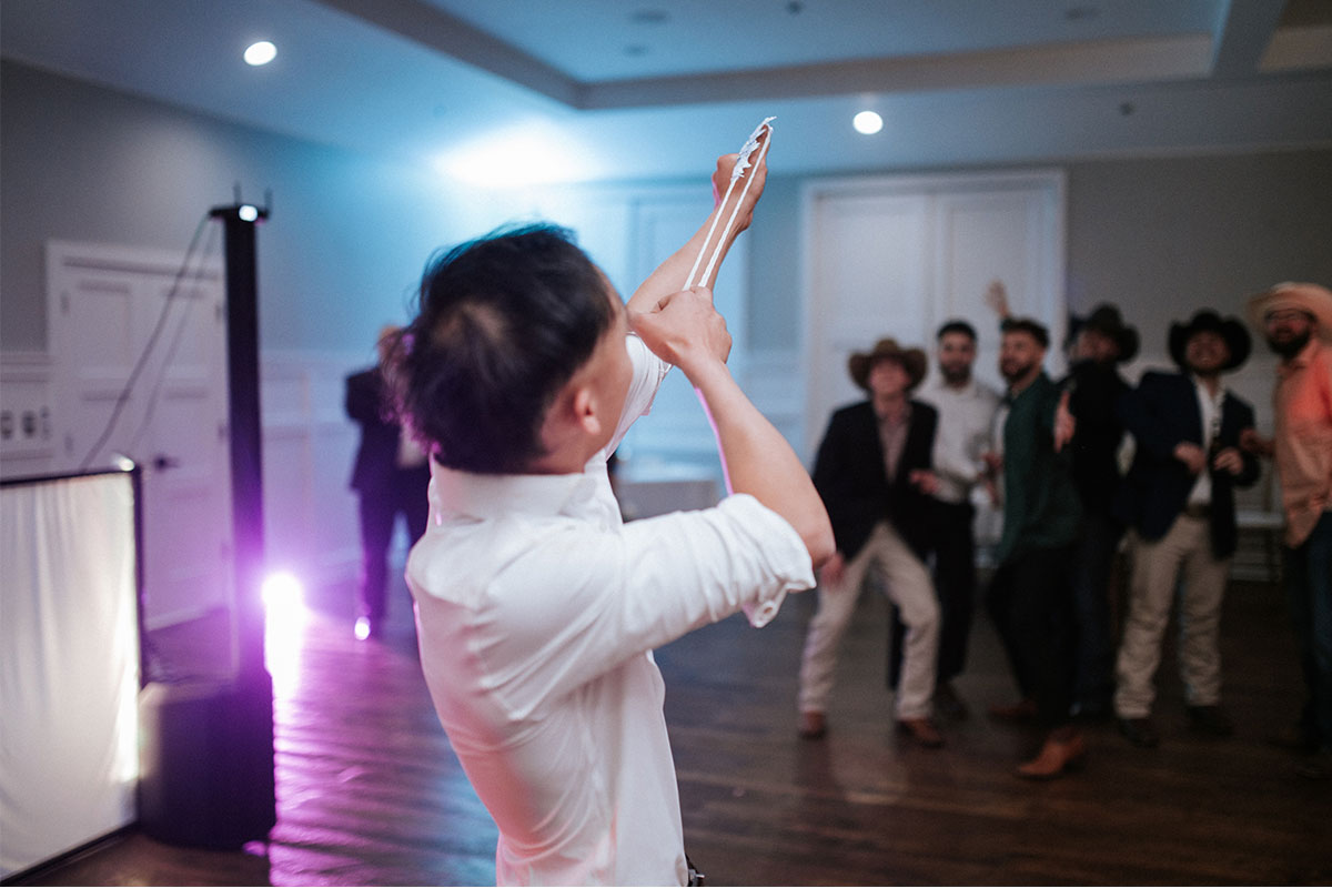 A man in a white shirt prepares to throw a garter as lively garter toss songs play, while a group of men in suits and hats stand ready to catch it in a decorated indoor event space.