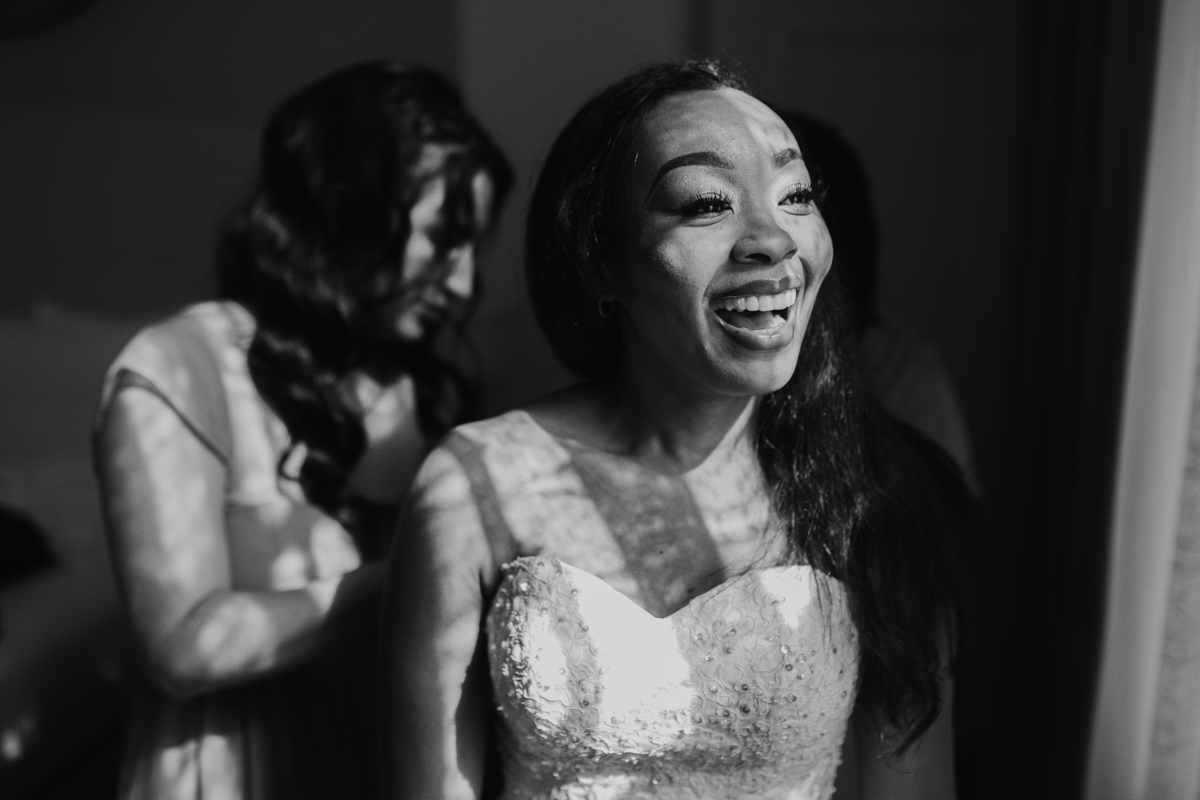 A woman in a wedding dress smiles brightly while another woman stands behind her, adjusting the dress, as Austin wedding photographers capture the joyful moment in a sunlit room.
