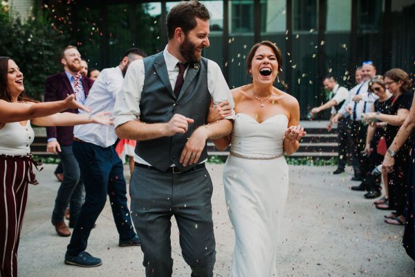 6-Benefits-Of-Hiring-A-Professional-Event-Videographer1 A bride and groom walk arm-in-arm outdoors, laughing as guests throw confetti around them during a wedding celebration.