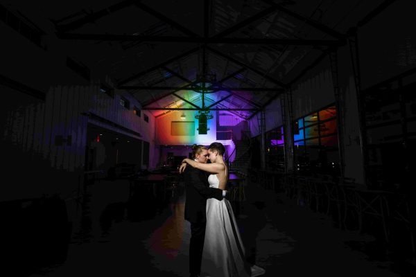 Brides-Dancing-min-scaled A bride and groom embrace while dancing in a dimly lit venue, illuminated by colorful, rainbow-like lights on the wall behind them.