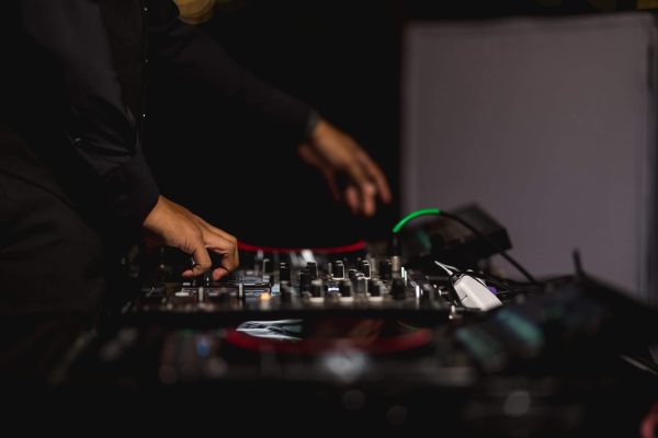 The-Importance-Of-Choosing-The-Right-Event-DJs-scaled-1 A person operating a DJ mixer and turntables with one hand adjusting controls and the other hand above the equipment, set against a dark background.