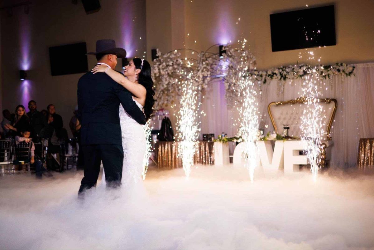 A couple dances in low fog as cold spark fountains fire behind them, with lit marquee letters and a floral backdrop.