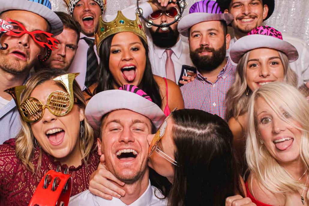 the-latest-wedding-photo-booth-trends-u-need-to-know A group of people wearing hats and novelty glasses pose closely together and smile at the camera; some stick their tongues out.