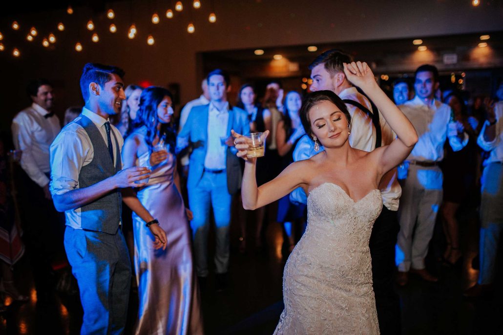 toast-wedding-DJ-bride-dancing-on-dance-floor A bride in a strapless wedding dress dances with a drink in hand at a reception, surrounded by guests on a dimly lit dance floor.