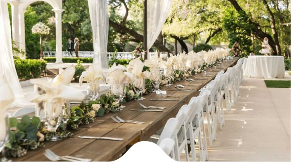 Event-Coordination-Planning-Services A long wooden table set with white flowers, glassware, and white chairs is arranged outdoors under a canopy, with trees and greenery in the background.