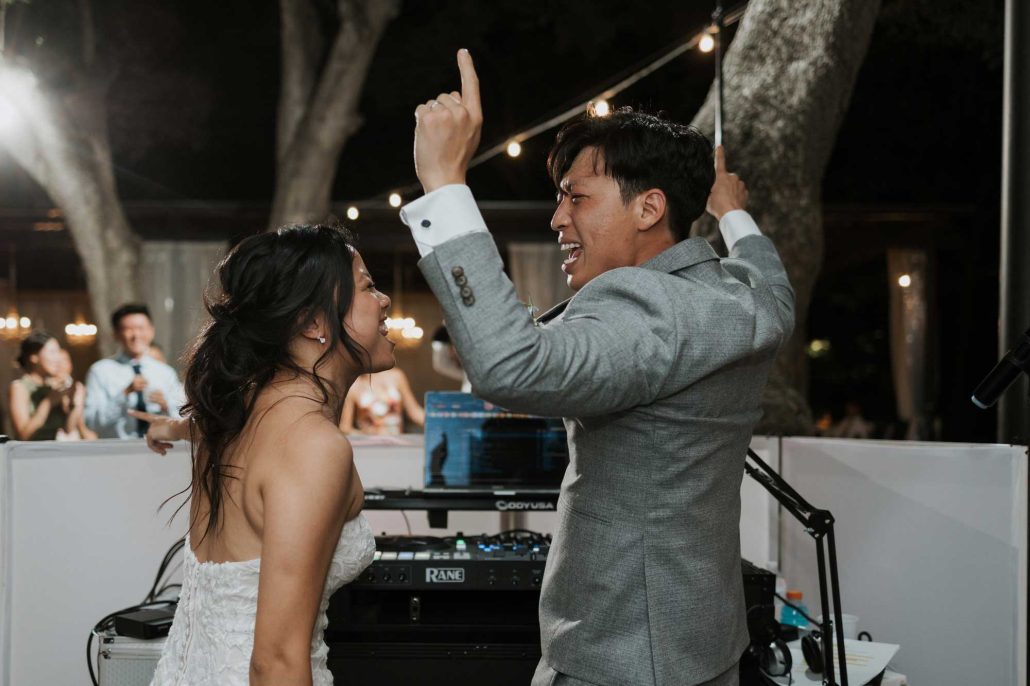 austin-dj-bride-groom-at-dj-booth A bride and groom in formal attire dance energetically near a DJ booth at an outdoor nighttime event with string lights.