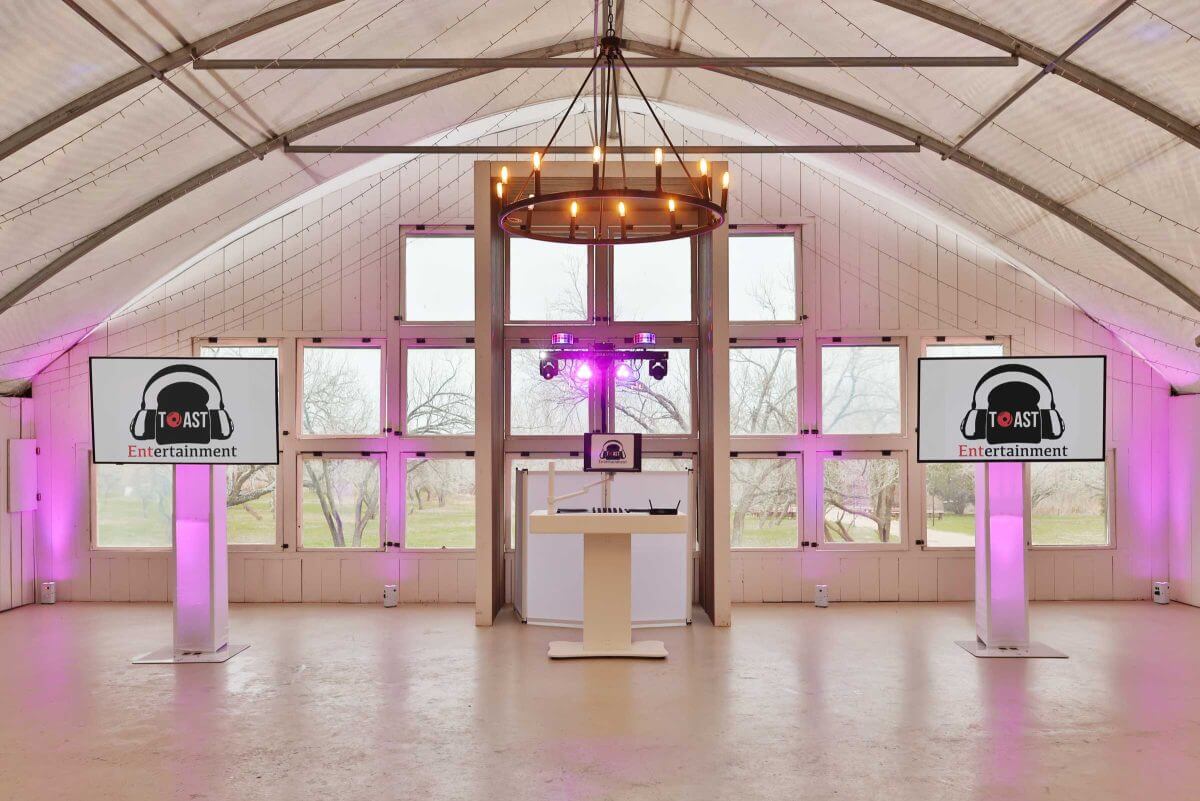 Two display screens on either side of a DJ booth in a barn venue lit with purple uplighting.
