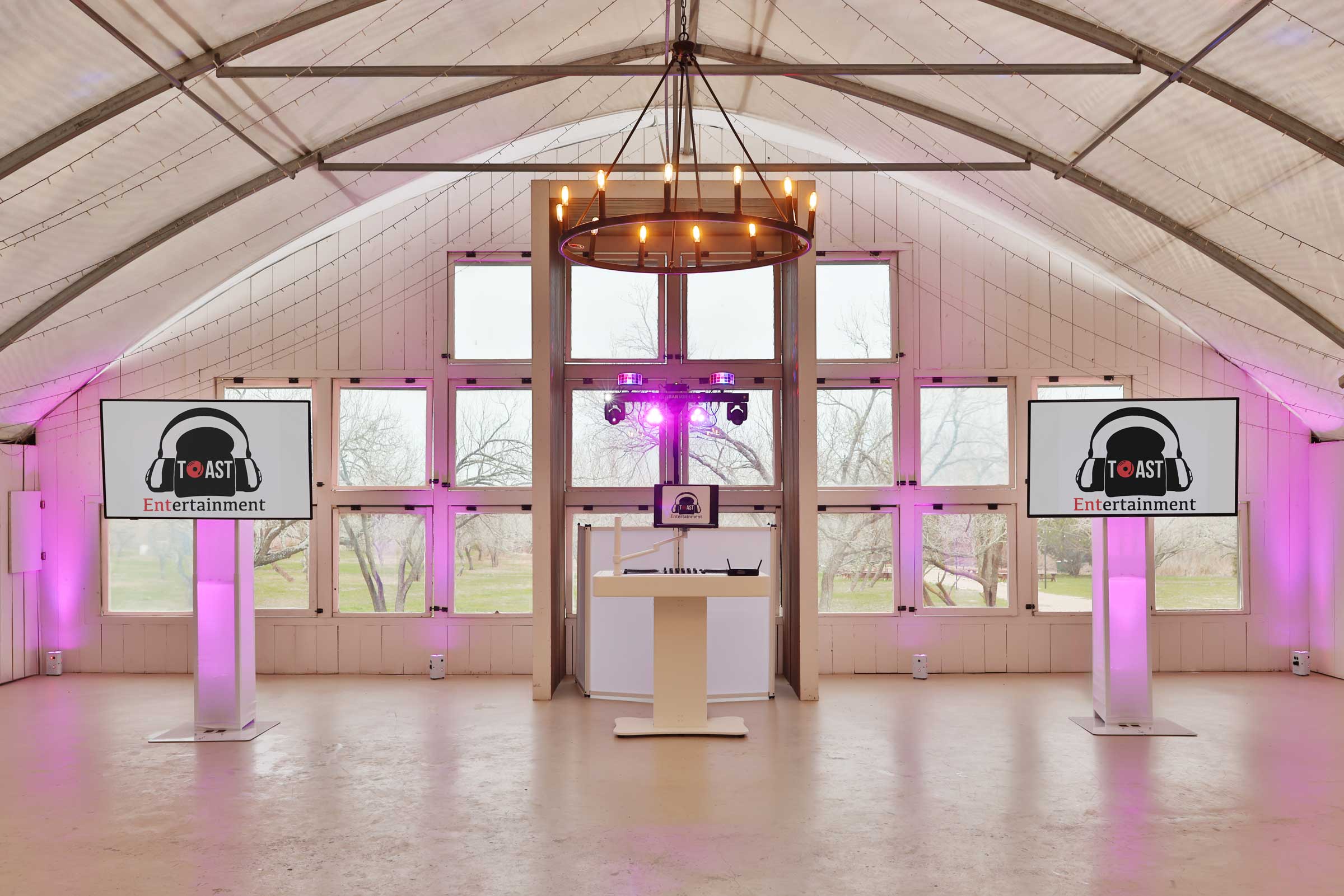 Toast-TV-Set-Up A modern event setup in a barn with two display screens showing "Toast Entertainment" logos, DJ booth in the center, and purple uplighting.