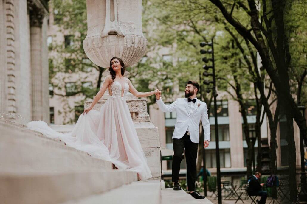 A bride in a white gown and a groom in a white jacket walk hand-in-hand outdoors on stone steps, with trees and buildings in the background.