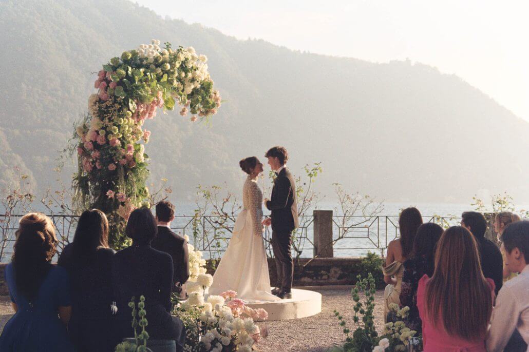 Houston wedding photography by Toast: a bride and groom stand under a floral arch during an outdoor wedding ceremony by a lake, with mountains in the background and guests seated nearby.