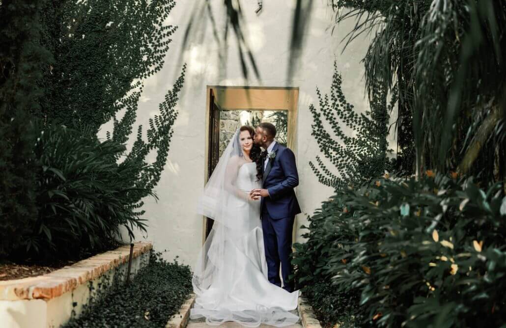 Houston wedding photography by Toast: a bride in a white gown and veil stands next to a groom in a navy suit, holding hands outside a building surrounded by greenery.