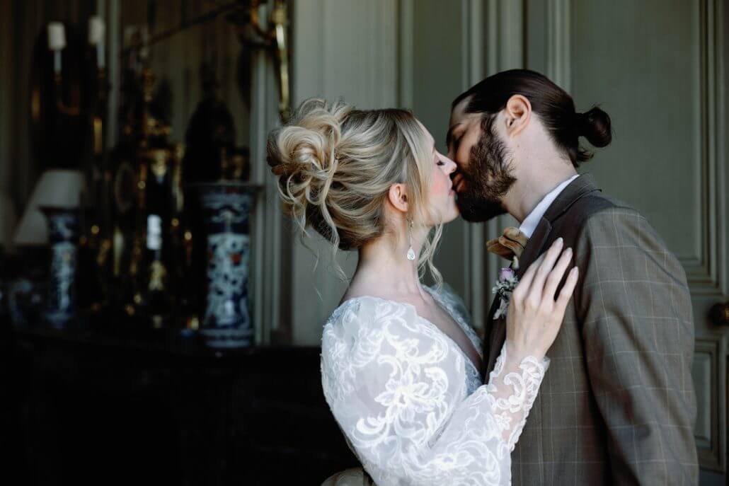 Houston wedding photography by Toast: a bride and groom share a kiss indoors, dressed in wedding attire. The bride wears a lace gown; the groom wears a brown suit. Ornate vases and decor are visible in the background.