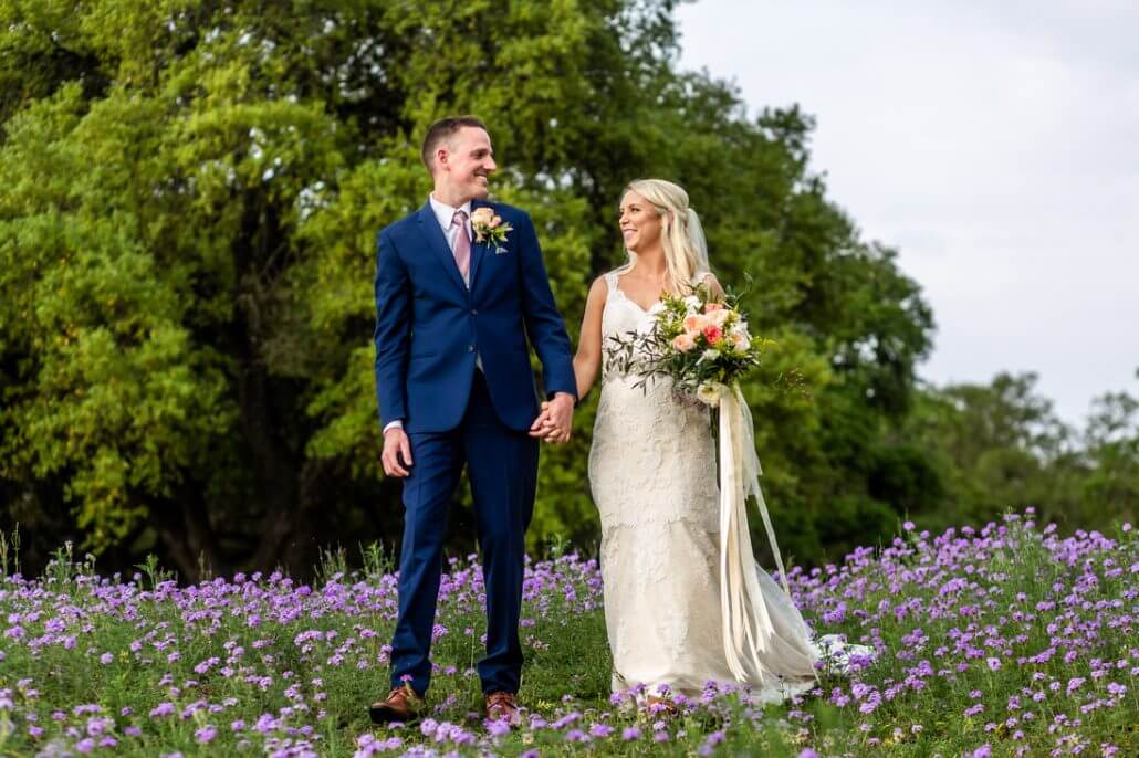 San Antonio wedding photography by Toast: a bride and groom hold hands and smile at each other while standing in a field of purple flowers, with green trees in the background.