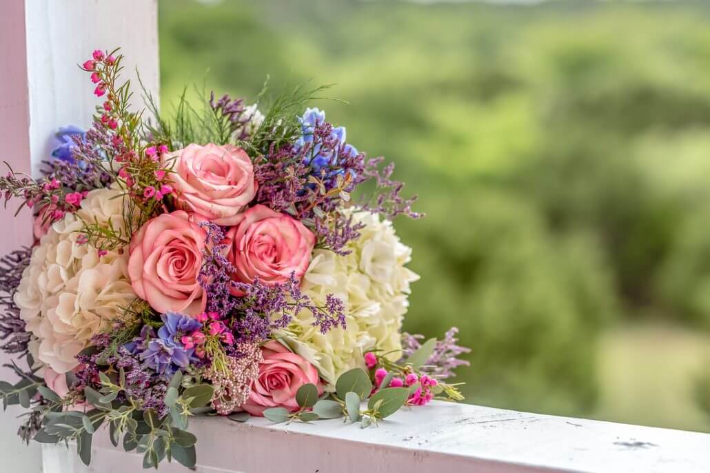 San Antonio wedding photography by Toast: a bouquet of pink roses, white hydrangeas, and purple flowers rests on a white wooden railing with a blurred green landscape in the background.